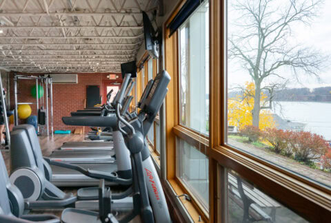 Indoor gym with treadmills and cardio machines near large windows with view of lake and autumn foliage.